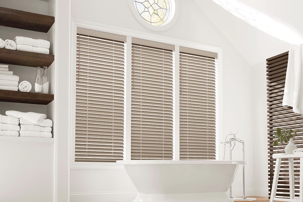 Three panels of custom made faux wood blinds filtering in light into a sleek bathroom with a beautiful stand alone tub.